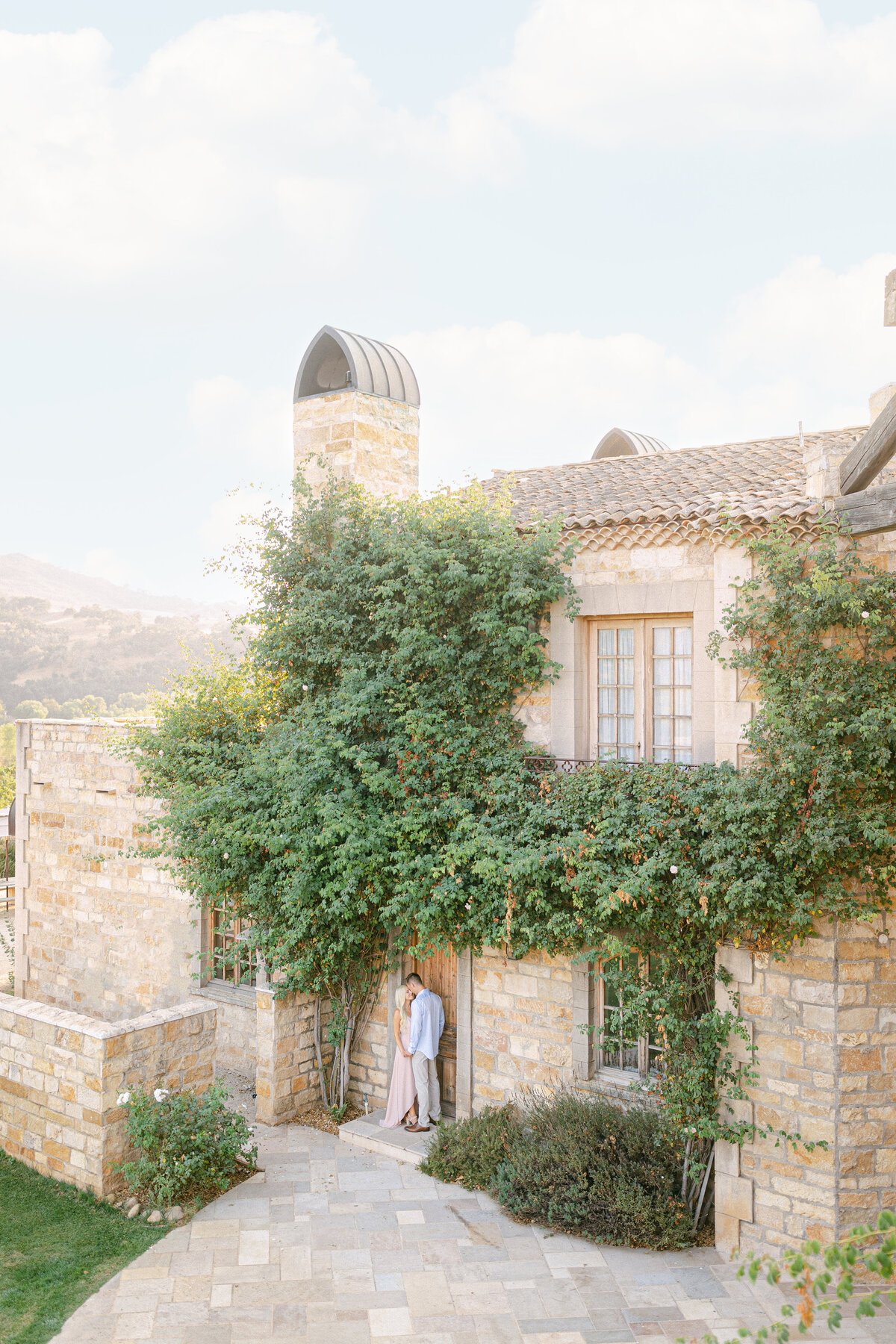 A kiss at the vine-covered door of the stone estate, hills gold behind