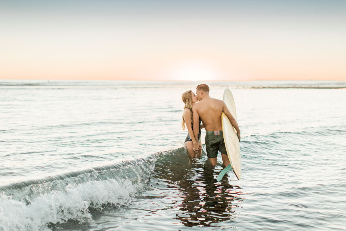 A kiss in the surf at sunset, his board under one arm