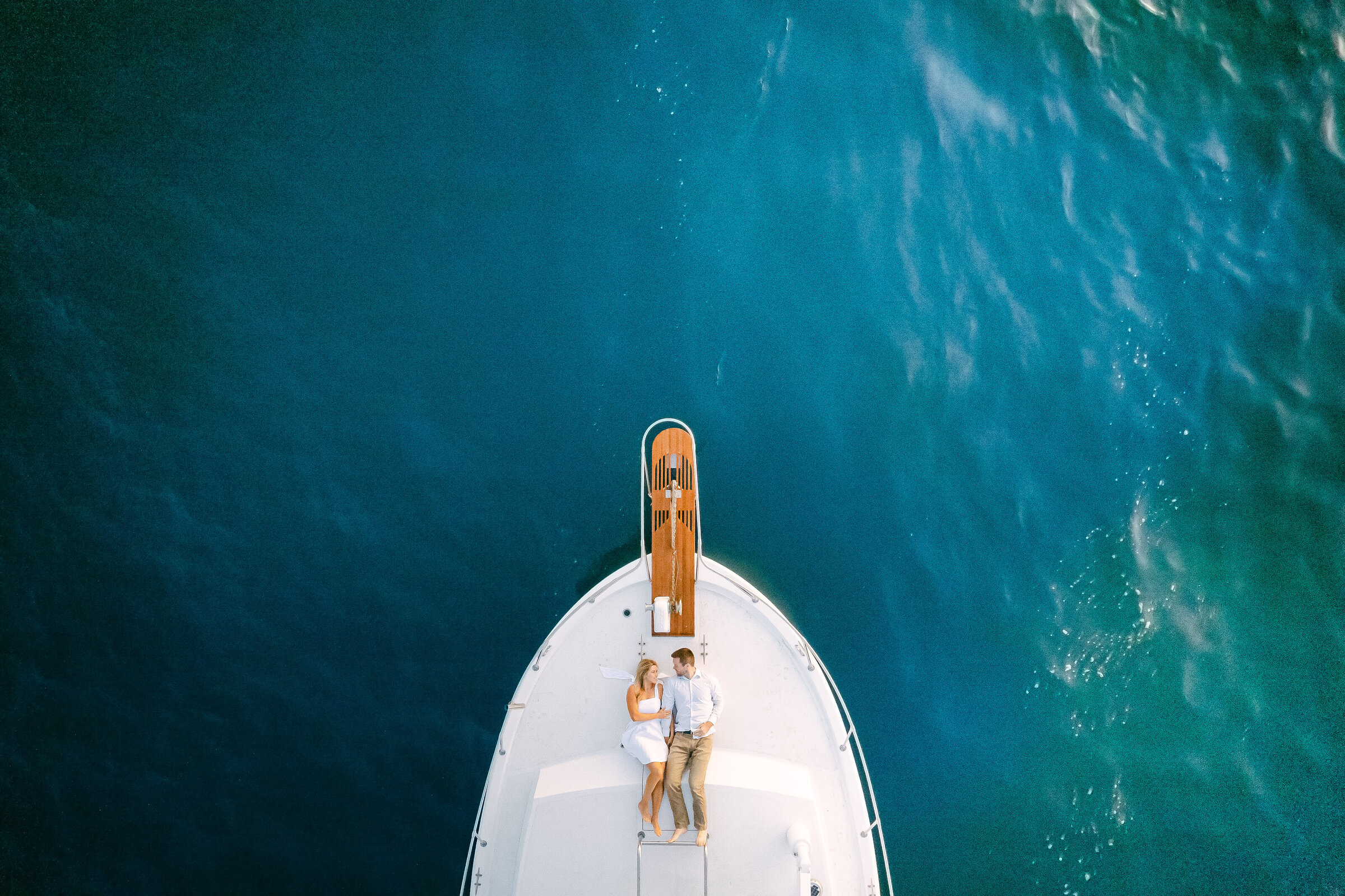 Aerial view of a couple lying together on the bow of a classic yacht over deep teal water